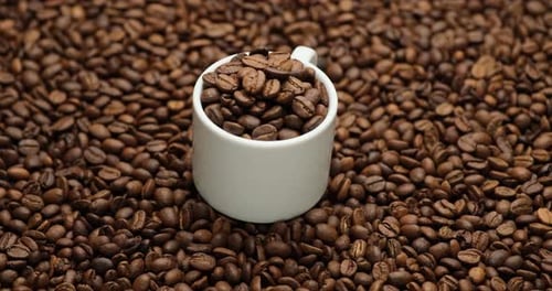 Keramiktasse voller Kaffeebohnen auf einem rotierenden Hintergrund aus frisch gerösteten Kaffeebohnen