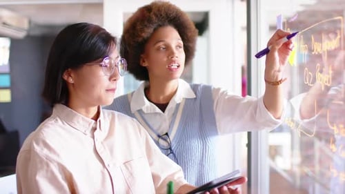 Duas colegas criativas diversas discutindo ideias em uma parede de vidro, em câmera lenta