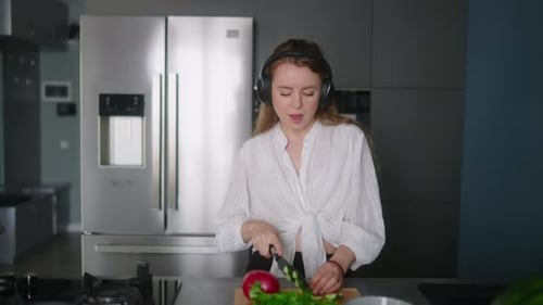 Woman in Headphones Making a Salad of Vegetables and Has Fun Dancing at Modern Kitchen Island Young