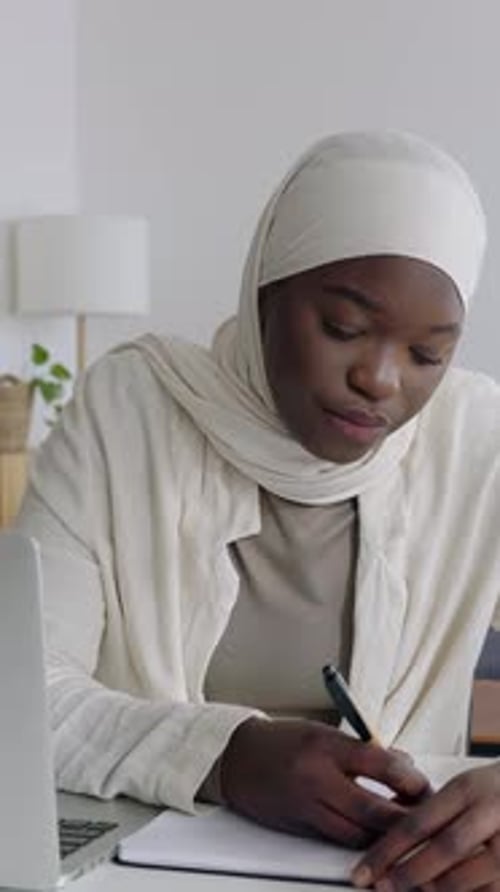 Junge afrikanische Studentin macht sich Notizen, während sie zu Hause am Laptop arbeitet