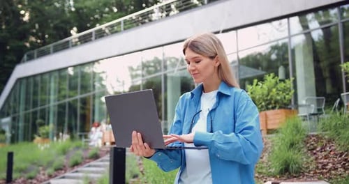 Smiling Businesswoman Typing on Laptop Outdoors Near Modern Office Building