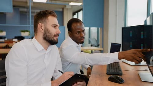 Zwei männliche Händler sitzen zusammen am Schreibtisch im Büro und überwachen Aktien, Daten, Kerzendiagramme auf dem Bildschirm