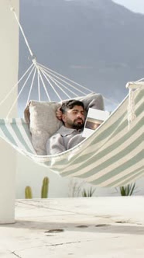 Vertical video: Reading man in hammock on roof with book closing eyes resting in light grey sweater