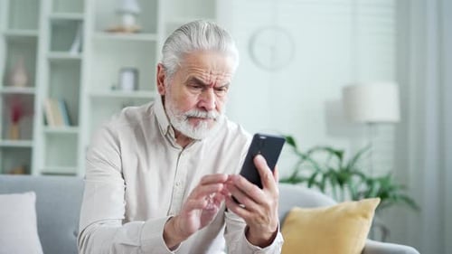 Frustrated elderly senior man having problems with software or service on smartphone sitting on sofa
