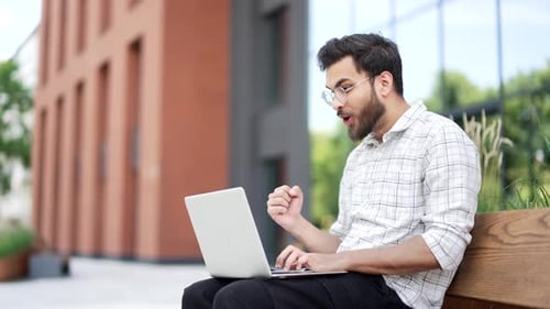 Satisfied happy handsome man received good news on laptop while sitting on a bench on the street