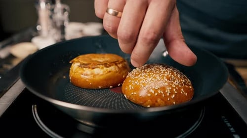 A close-up of a chef toasting a bun on a griddle while cooking a burger in a restaurant kitchen