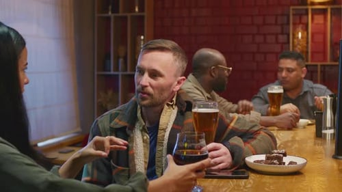 Man Speaking with Lady at Bar Sitting with Glasses of Beer and Wine