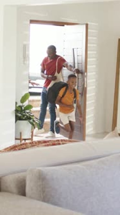 Un père afro-américain et son fils adolescent entrent dans un salon lumineux, transportant un sac à dos et des produits