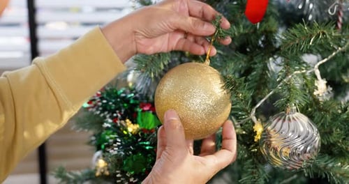 Close-up of hands placing a golden ornament on Christmas tree. Person decorates tree with glittery