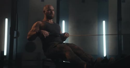 Bearded Muscular Man in a Tank Top Working Out on a Rowing Machine in a Modern Gym