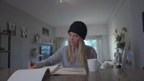 A beautiful blonde woman writing on a notebook and drinking a hot beverage at home