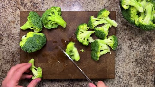 A head of broccoli is separated into florets using a knife against a wooden board.