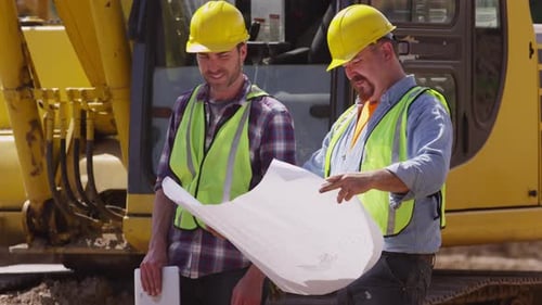 Construction workers review blueprints by heavy excavation equipment