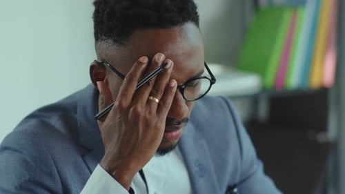 Face: un joven afroamericano cansado se quita las gafas en la oficina, sentado en el escritorio, proyecto de dolor de cabeza