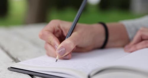female hands writing in journal, sitting on white table in the green garden, handheld closeup