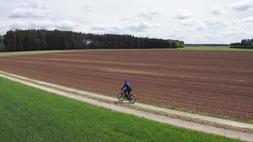 Aerial wide shot of a man riding a bike on a small dirt road. Fields in the foreground, forest in th