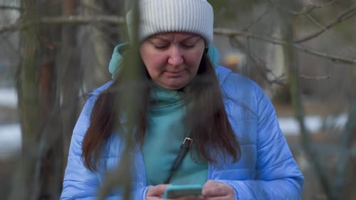 Middle Aged Woman Use Smartphone Outdoors in Park Standing Behind Tree Branches