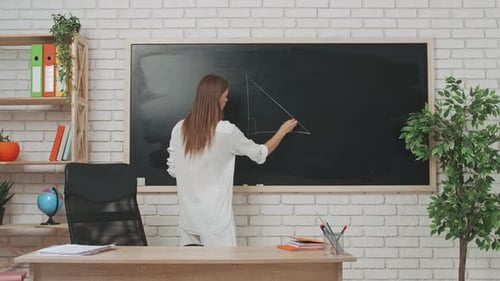 Young Woman College Teacher at Desk in Classroom in Front of Chalkboard Explains Geometry Lesson to