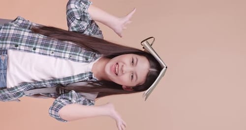 Cute school girl is carrying a school bag and holding a notebook on her head on brown background.