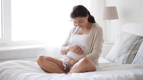 Pregnant woman holding belly, sitting in bright bedroom