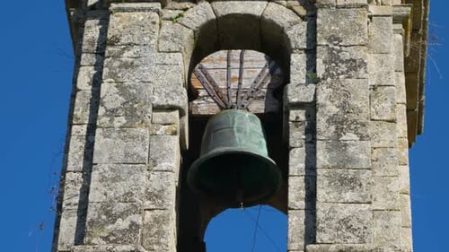 Pan across church bell suspended in old weathered stone cut tower fronting blue sky