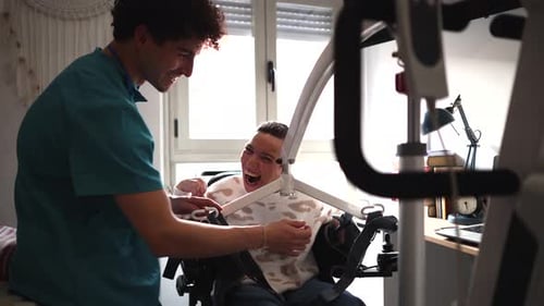 Young male caregiver assisting a laughing disabled woman in a wheelchair, using a patient hoist lift