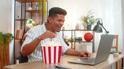 Indian Man Enjoys Popcorn Laughs at Comedy Film on Laptop Watches Funny Scene Smiles Relax at Desk