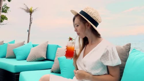 Beautiful Young Woman in a Straw Hat Lounges on a Beach Couch Enjoying a Tropical Cocktail