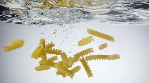 Mix of pasta noodles thrown into clear water with nice bubbles, slow motion, close up
