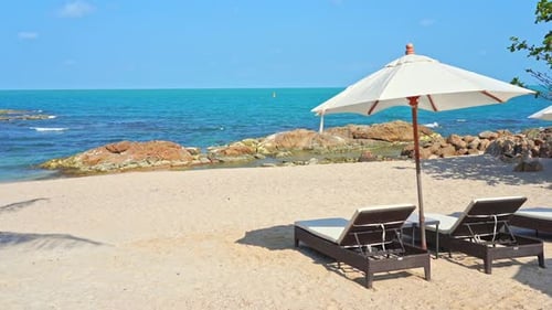 Two empty sun loungers under a sun umbrella facing a picturesque sandy beach and ocean view. Title s