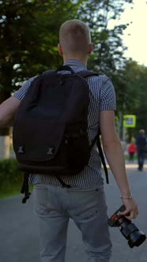 Vertical of A Student Photographer is Walking Along a City Street Behind Him