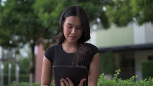 Portrait of beautiful young Asian Thai woman using technology outdoors while sitting in park alone