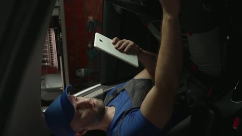 Vertikales Video: Ein Mechaniker mit blauem T-Shirt und Kappe sitzt im Auto und führt Diagnosen durch