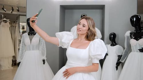 Retrato de noiva loira fazendo selfie com foto usando smartphone enquanto experimenta o vestido no salão de casamento