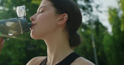 Caucasian girl drink clear mineral water after jogging training in city park.
