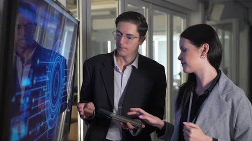 Two people are discussing cybersecurity in an office, interact with a digital screen showing data.