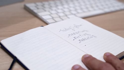 Close-up hand writing today's marketing task list in notebook on modern office desk workspace