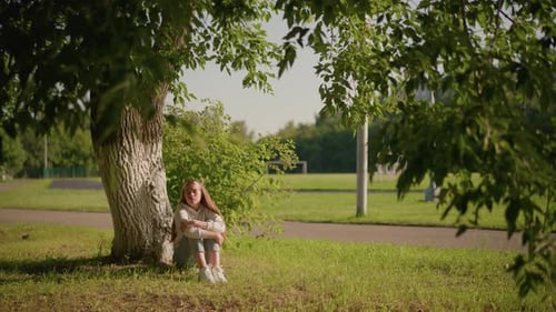 Thoughtful Woman Sitting Under Tree in Sunlit Park with Stadium Background