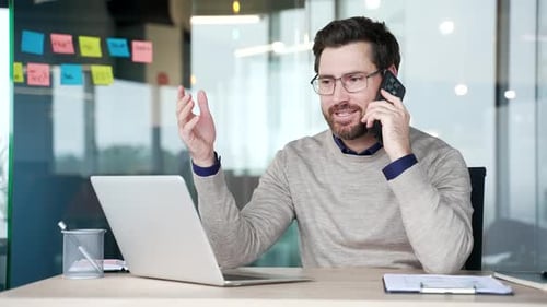 Businessman talking on smartphone while sitting at desk at workplace in modern glass office. Mature