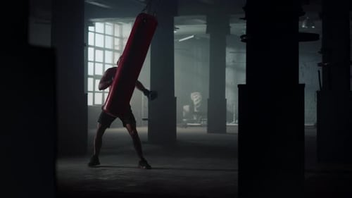 Aggressive african american boxer silhouette training hard with punch bag in loft gym