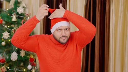 Man Wearing Santa Hat Adjusts Clothes by Christmas Tree