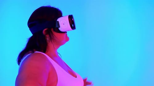 Woman with overweight in a tracksuit and VR headset in a studio with blue background and red illumin