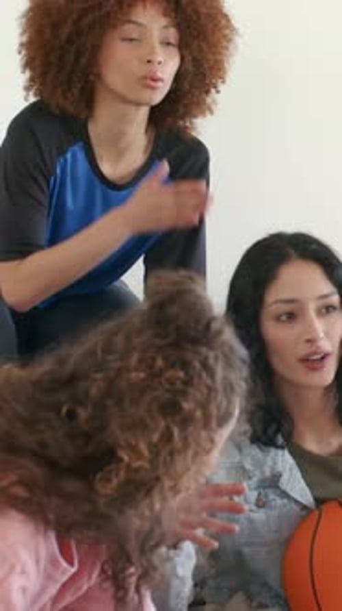 Vertical video: Reacting trio to TV on couch, pink knit leaning, holding glass, orange basketball