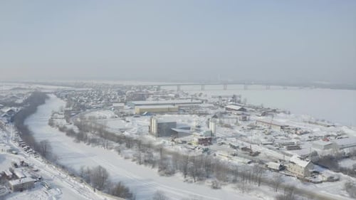 Luftbild einer gefrorenen Kleinstadt in der Wintersaison Clip, der über einem verschneiten und eisbedeckten Fluss fliegt und