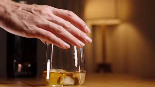 A woman's hand shakes a glass with whiskey and ice and places it on the wooden table, close-up. Slow
