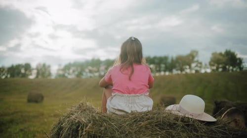 Rear View of Girl Sitting on Hay Bale While Boy Focuses on Hay in Open Field