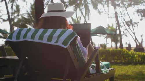 Freelancerin arbeitet am Laptop und sitzt in der Sonnenliege im tropischen Resort, Rückansicht