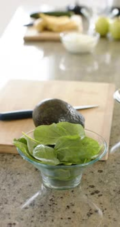 Vertical video: Camera is holding focus on glass bowl of greens on stone counter, showing prep