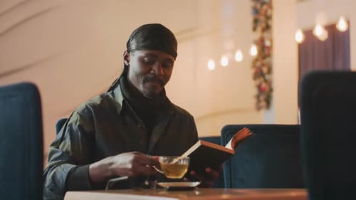 Black Man Traveler Smiling While Reading and Sipping Tea in Cozy Cafe Warm Decor Relaxed Posture