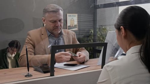 Customer Handing Money to Bank Teller Across Glass Counter in Bank